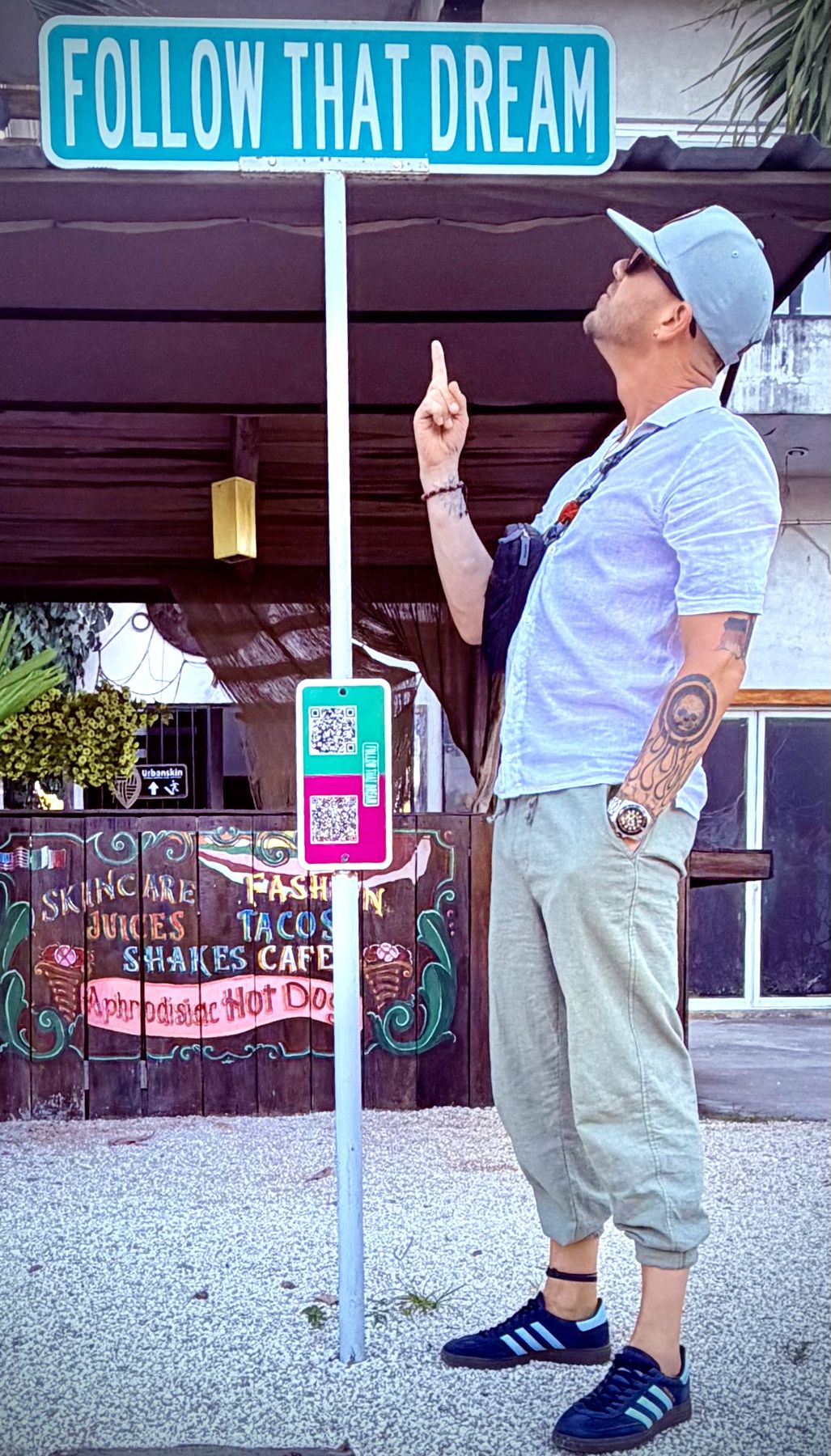Timothy pointing up at a teal and white street sign that reads Follow That Dream, beside a painted cafe fence in the Yucatan